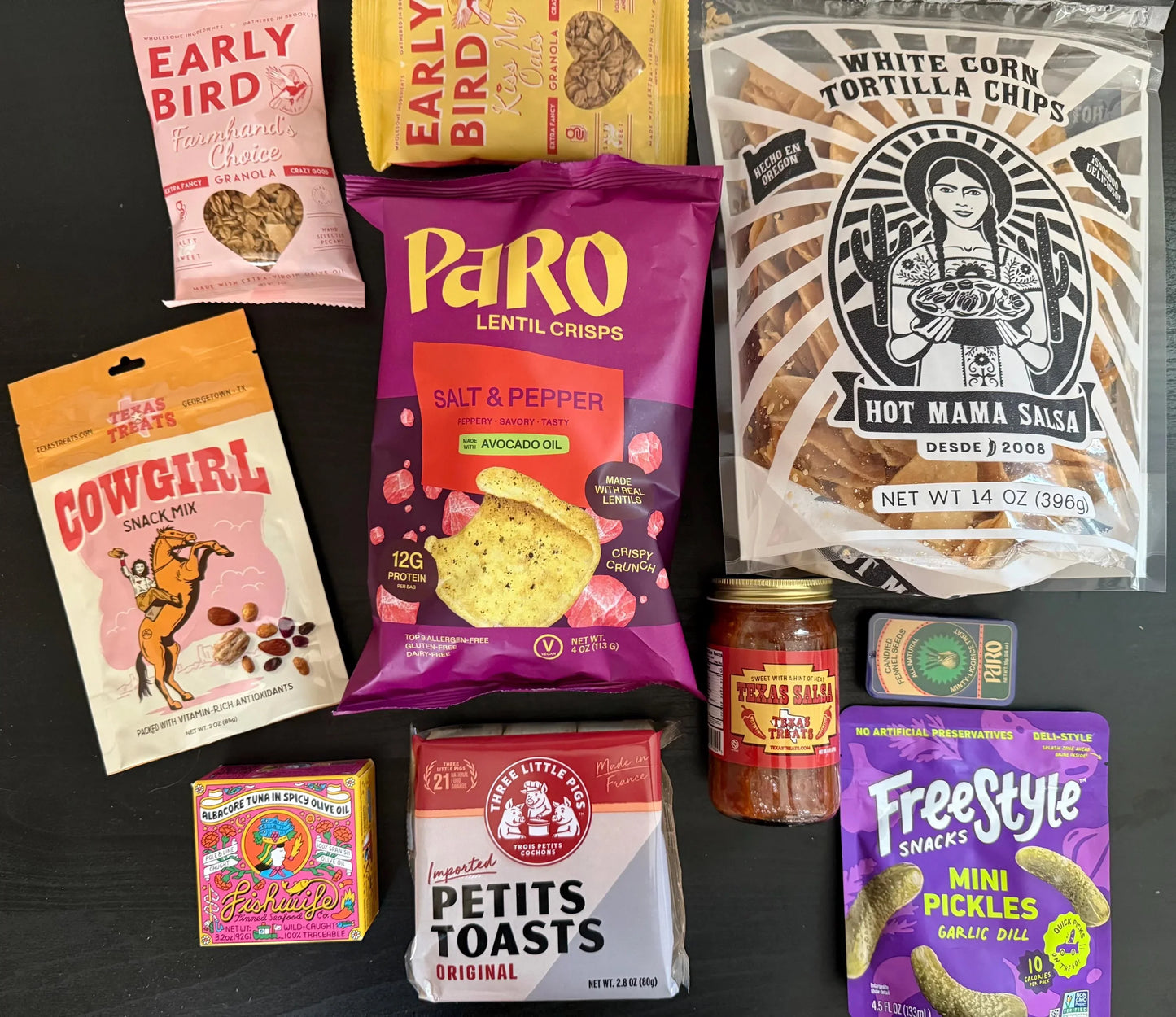 Assorted gourmet snack foods including granola, lentil crisps, tortilla chips, snack mix, salsa, toasts, pickles, and tinned fish on a dark surface.