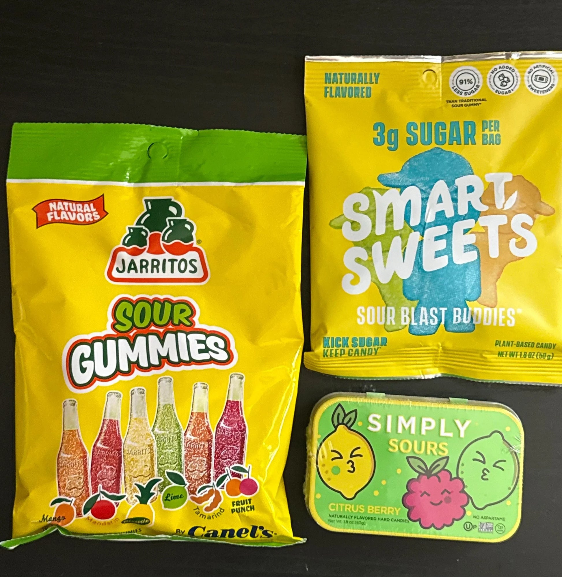 Assorted sour candy packs including Jarritos sour gummies, SmartSweets Sour Blast Buddies, and Simply Sours citrus berry tin on a dark surface.