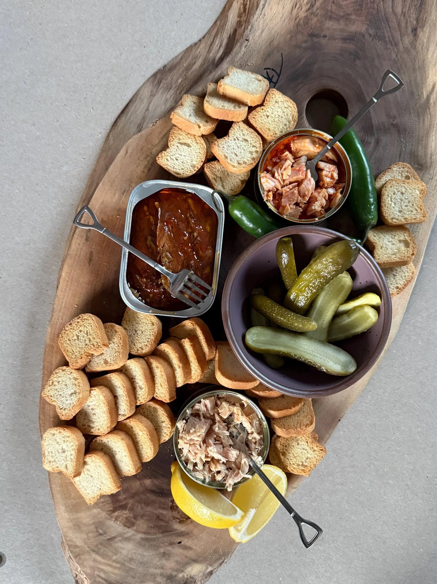 Charcuterie board with canned fish, pickles, crostini, lemon wedges, and peppers