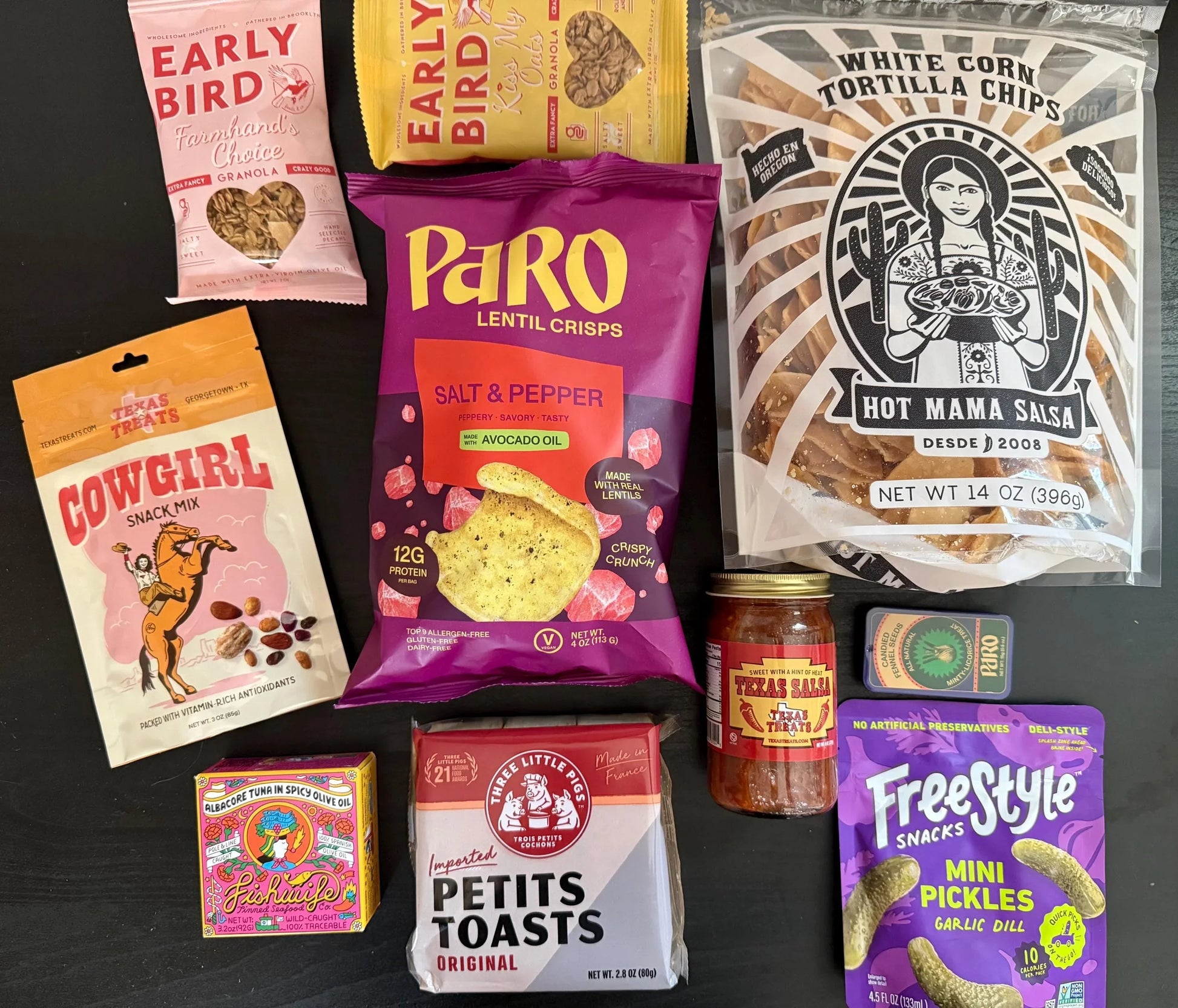 Assorted gourmet snacks and artisan foods including chips, granola, salsa, and pickles on a black background.