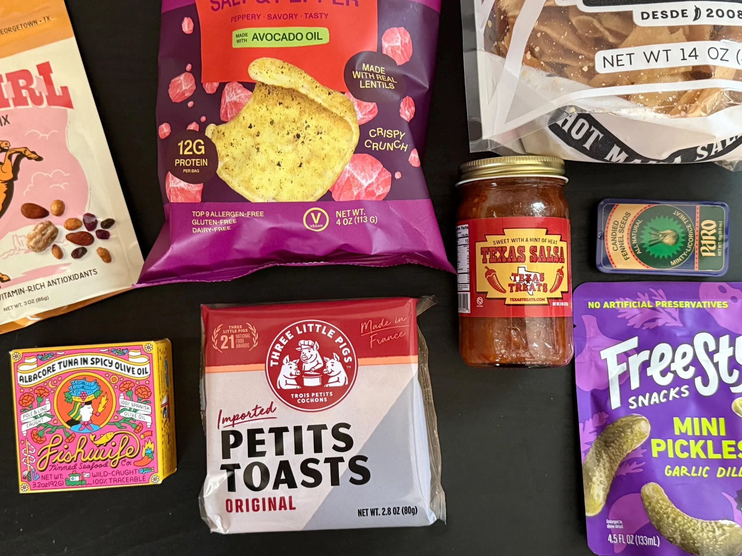 Assorted gourmet snacks, salsa, pickles, tinned tuna, and crisps on a black surface.
