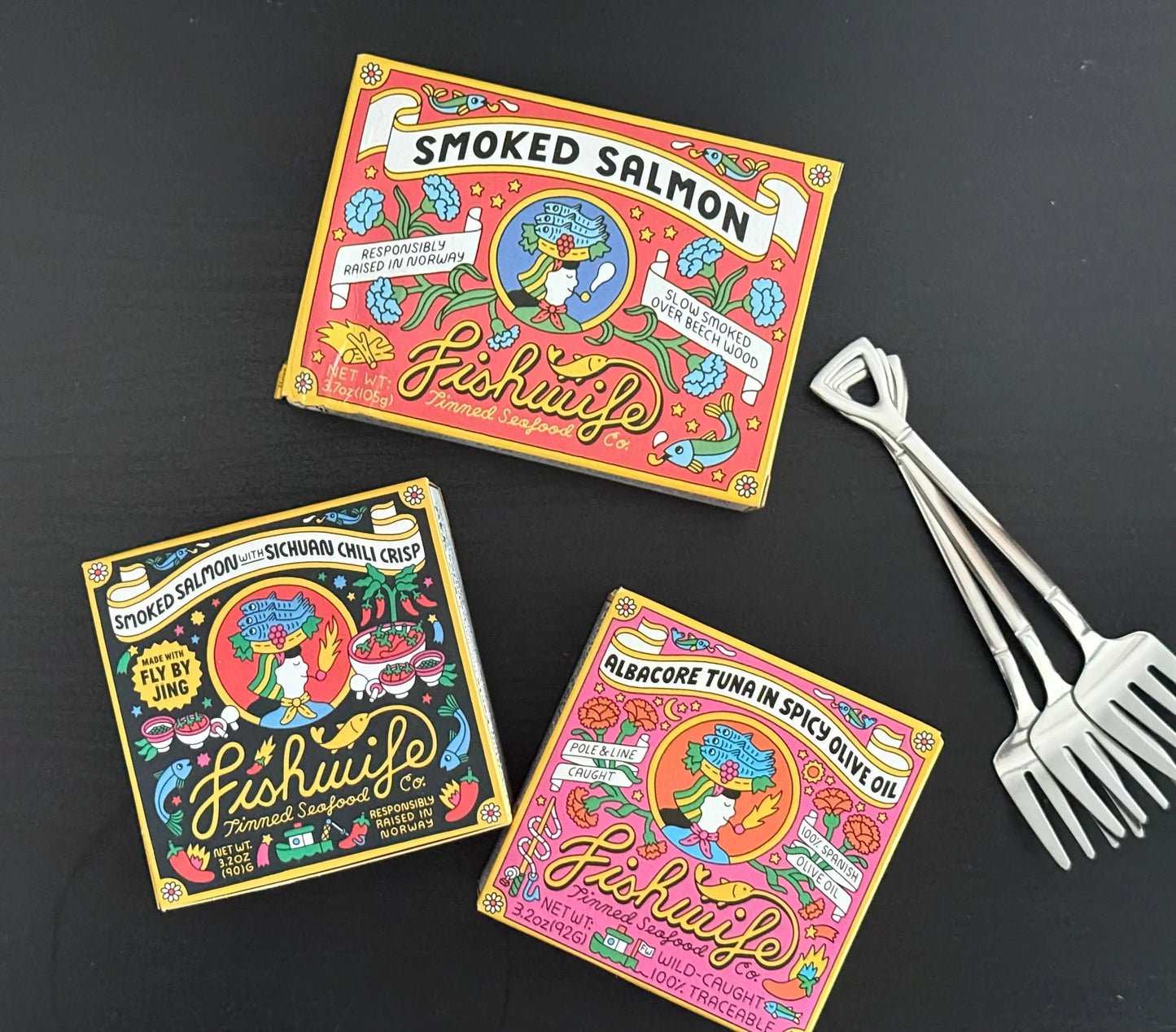 Three colorful tins of Fishwife smoked salmon and tuna on a black table with metal seafood forks.
