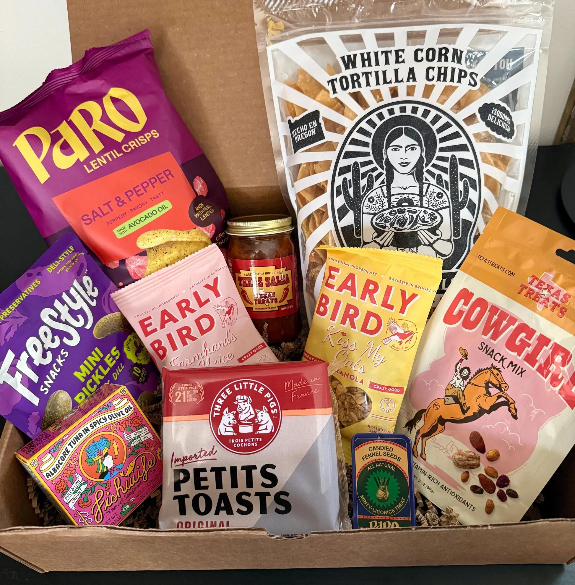 Assorted gourmet snacks and treats in a gift box, including chips, crisps, pickles, and granola.